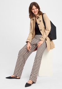Beige trenchcoat met zwarte knopen, gestreepte broek in crème en bruine golven, en zwarte puntige ballerina's. Inclusief een zwarte tote bag.