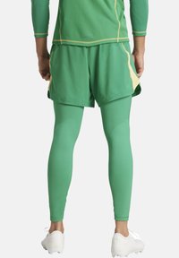 Abbigliamento sportivo verde composto da una maglietta a maniche lunghe in rete, pantaloni corti abbinati con un accento giallo e leggings verdi aderenti. Scarpe bianche.