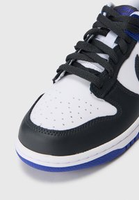 Czarno-białe sneakersy z szarymi sznurowadłami, posiadające perforowaną część palcową oraz niebieskie akcenty na języku i podeszwie. Gładka skórzana powierzchnia.