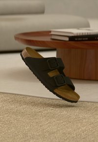 Μαύρη σαγιονάρα με δύο λουριά και ρυθμιζόμενες αγκράφες, με πάτο από φελλό, αιωρείται πάνω από μπεζ χαλί κοντά σε ξύλινο τραπεζάκι σαλονιού και λευκό καναπέ.