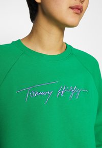 Sweatshirt vert avec un col ras du cou côtelé et un logo signature orné de strass sur la poitrine. Tissu doux et texturé.