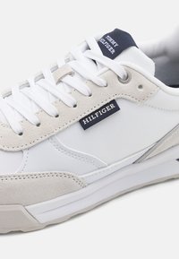 Hvit sneaker med en kombinasjon av syntetiske materialer og semsket skinn, med et teksturert design, hvite snørebånd og en marineblå merkeetikett.