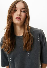 Camiseta de manga corta gris oscuro con un ajuste relajado, que presenta piedras de strass decorativas dispuestas en líneas verticales en el frente.