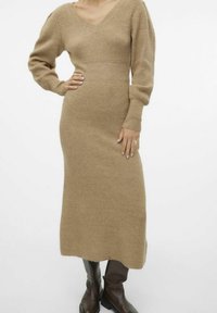 Abito a maglia a maniche lunghe di un morbido beige con scollatura a V, texture a costine, vestibilità a sigaretta e lunghezza a metà polpaccio; abbinato a stivali marrone scuro.