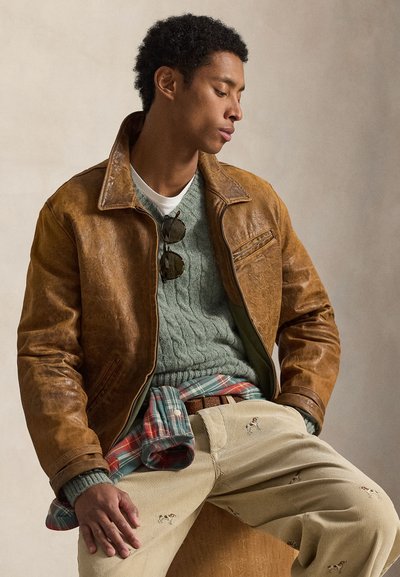 Καφέ δερμάτινο μπουφάν με γιακά, φορεμένο πάνω από πράσινο πουλόβερ με πλεκτό σχέδιο, σε συνδυασμό με μπεζ παντελόνι κορντόρι με κέντημα αλόγου.