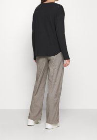 Kobieta w czarnej bluzce z długim rękawem, luźnych beżowych spodniach w kratę i białych trampkach, stojąca na tle jednolitego białego tła, tyłem do obiektywu.