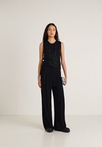 Črno brezrokavnik z reliefnimi detajli, kombiniran s širokimi črnimi hlačami in temnimi čevlji. Model drži majhno srebrno torbico.