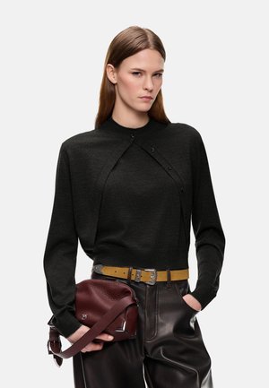 Donna con capelli lisci castano chiaro indossa un top nero a strati, cintura senape, pantaloni di pelle scura e tiene una borsa a mano in pelle rosso scuro.