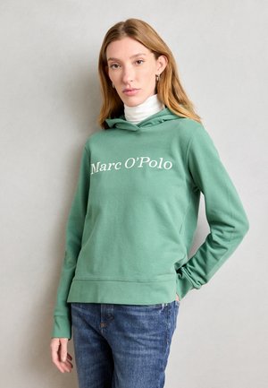 Sudadera verde hecha de tejido suave con un bolsillo tipo canguro, que presenta un texto de logo blanco "Marc O'Polo" y puños acanalados. Combinada con vaqueros azules.