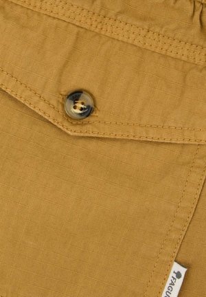 Gros plan sur une poche en tissu jaune moutarde avec un bouton rond marron et une petite étiquette blanche portant la mention « FAGUO » sur le bord.