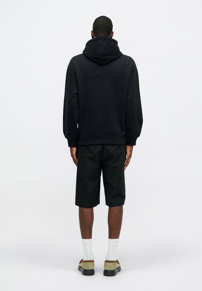 Sweatshirt à capuche noir avec une coupe décontractée, associé à un short vert foncé. Tissu texturé, poignets côtelés et sandales plates avec une semelle légère.