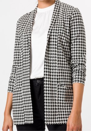 Femme portant un blazer ouvert à motif pied-de-poule noir et blanc, une blouse blanche et un pantalon noir, debout devant un fond uni.