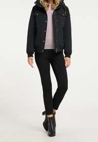 Giacca nera isolata con chiusura a zip e bottoni, due tasche sul petto e maniche con polsini, abbinata a pantaloni neri aderenti e stivaletti alla caviglia.