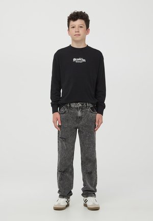 T-shirt nera a maniche lunghe con grafica "Brooklyn", abbinata a jeans grigi sbiaditi con un motivo strappato. Sneakers bianche e nere.