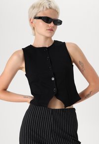 Top court sans manches noir avec un col rond, fermeture à boutons à l'avant et ourlet incurvé. Fabriqué en tissu lisse avec un design ajusté.