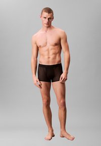 Czarne boxerki z wzorzystym wzorem, z wygodną gumką w pasie. Model stoi boso na jednolitym szarym tle.