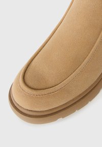 Acercamiento de la punta de una bota de ante color beige con forma redondeada, costuras detalladas y suela gruesa marrón sobre un fondo blanco.