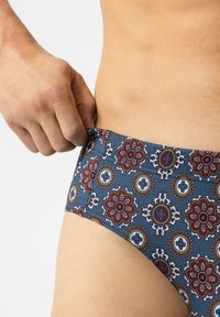 Blauwe zwemslip met cirkelvormige bloemmotieven in rood en wit. Het materiaal lijkt soepel met een elastische tailleband. Hand die aan de zijkant aanpast.