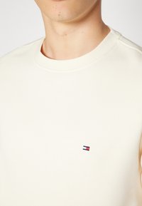 Camiseta de algodón crema de cuello redondo con un pequeño logo bordado en rojo, blanco y azul en el lado izquierdo del pecho. Textura suave.