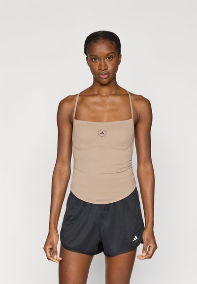 Débardeur beige moulant avec fines bretelles et encolure arrondie, décoré d'un logo circulaire. Associé à un short de sport noir.