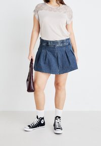 Ljusbeige topp med spetsdetaljer, denimkjol med fällningar och svarta högstövlar. Håller en liten handväska i vinröd färg.