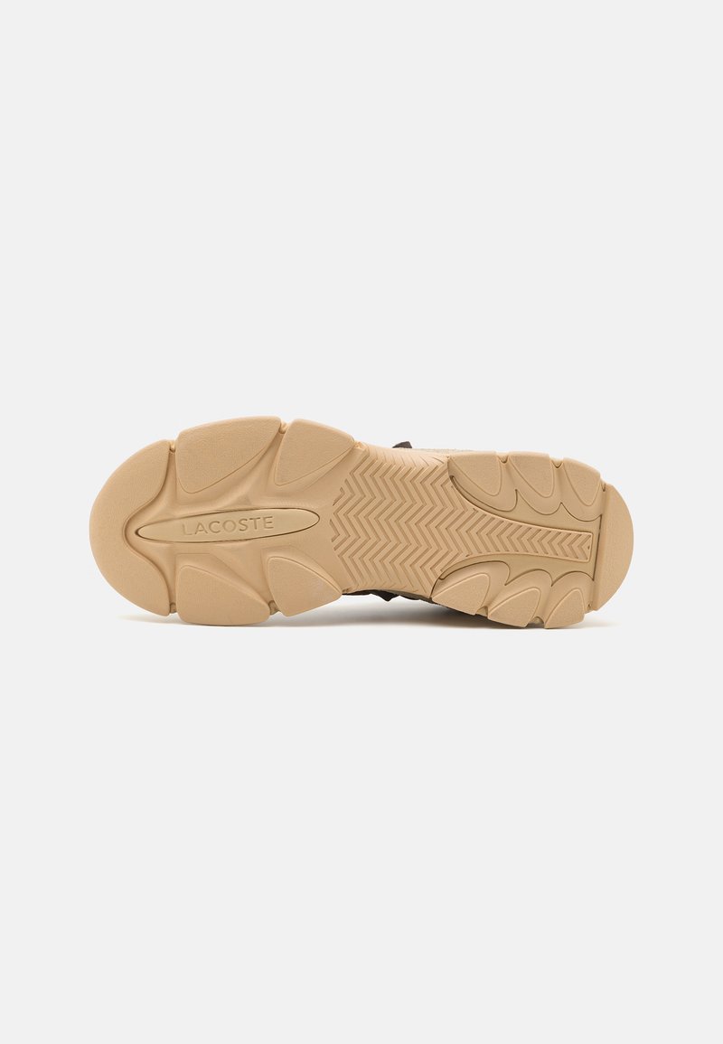 Czarna podeszwa sneakersów z teksturowaną, jasnobrązową gumową podeszwą zewnętrzną, z wzorem w zygzak oraz wytłoczonym na środku logo "LACOSTE".