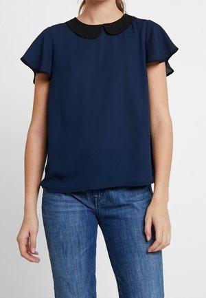 Person trägt eine marineblaue Bluse mit flatternden Ärmeln und einem schwarzen Peter-Pan-Kragen, kombiniert mit blauen Jeans vor einfachem Hintergrund.