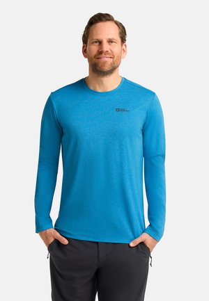 Man in een felblauw lange mouwen shirt met een klein logo op de borst, zwarte broek, staand met handen in de zakken tegen een witte achtergrond.