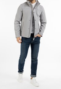 Veste softshell grise avec un col haut, des boutons-pression et des poches latérales, associée à un jean foncé et des baskets blanches.