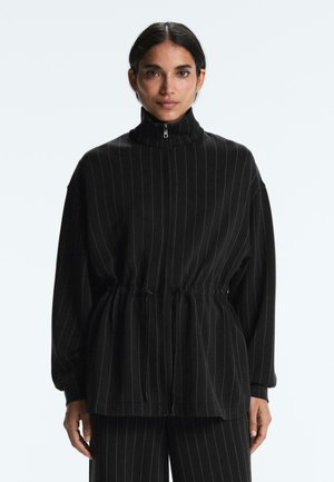 Ženska, oblečena v črno jopa z razčlenjenimi belimi črtami, z visokim ovratnikom in srajčnim pasom z vrvico, v kombinaciji z ujemajočimi hlačami z belimi črtami, na belem ozadju.