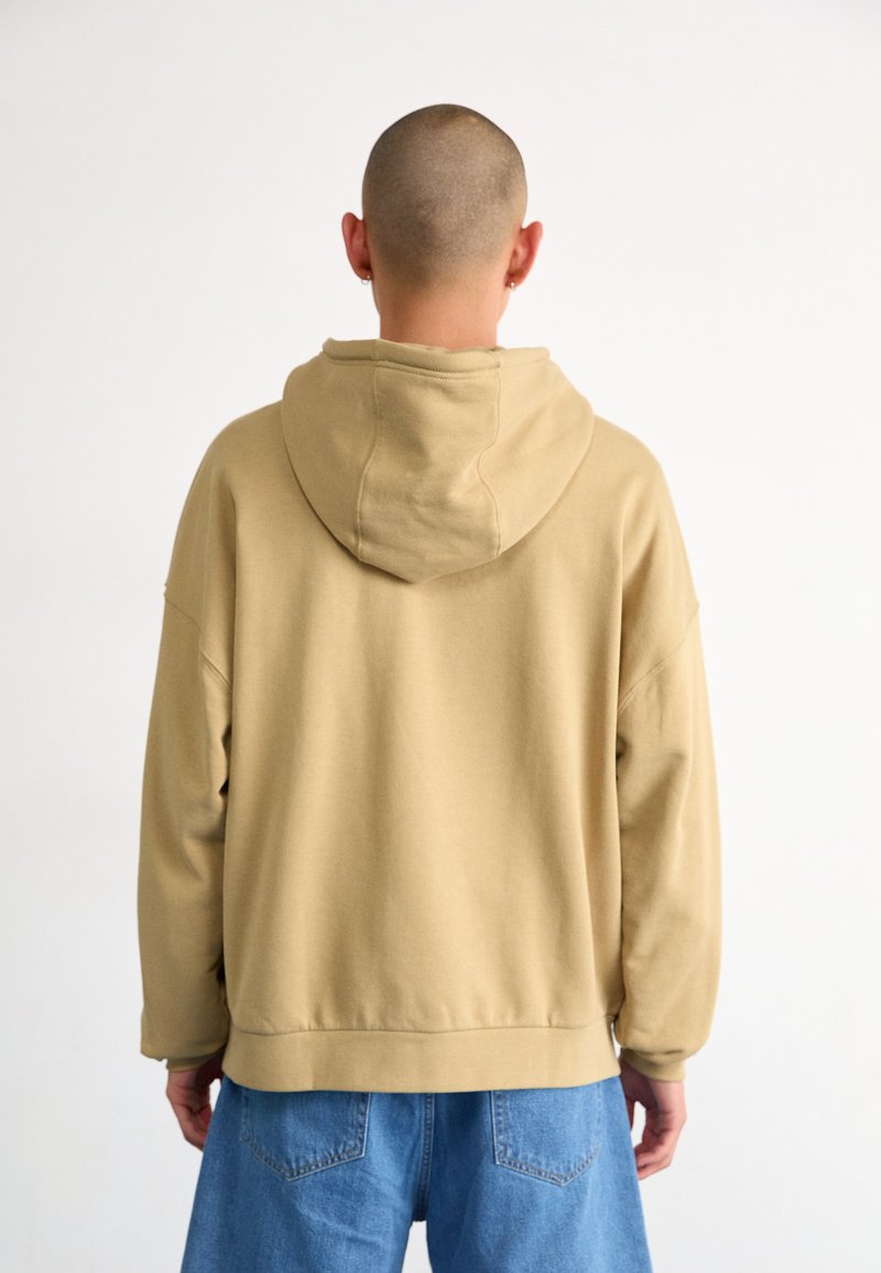 Haut à capuche beige oversize avec un cordon de serrage et un ourlet côtelé, fabriqué en tissu doux, porté sur un jean bleu clair ample.