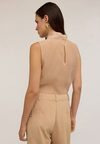 Top beige senza maniche con colletto alto e dettaglio a chiave sul retro, abbinato a pantaloni beige su misura con tasche laterali e tessuto strutturato.