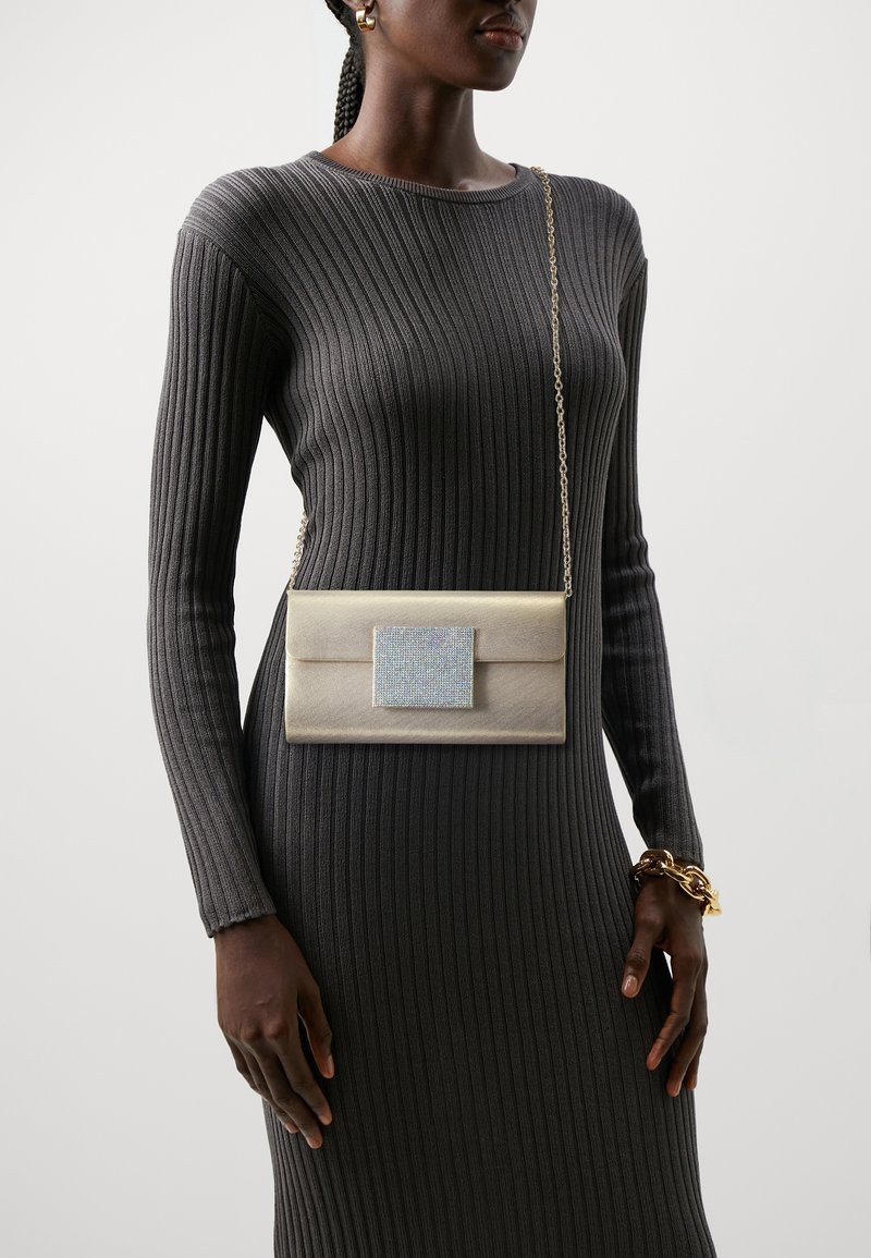 Femme portant une robe côtelée foncée avec un sac bandoulière beige doté d'une grande boucle carrée en strass et d'une bandoulière chaîne dorée.