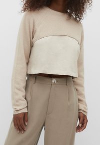 Beżowy sweter krótkim rękawem o dzianinowej teksturze i długich rękawach, nałożony na jasnobeżową bluzkę, w zestawie z beżowymi spodniami z detalem guzika.