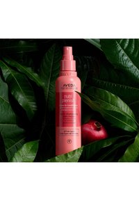 Le revitalisant sans rinçage Aveda Nutri Plenish dans un flacon en spray rose, avec un bouchon pompe, placé parmi des feuilles vert foncé et une grenade.
