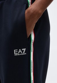 Χέρι που ακουμπά στην τσέπη σκούρου μπλε παντελονιού Emporio Armani EA7 με πλευρική ρίγα σε λευκό, πράσινο και κόκκινο χρώμα με λογότυπο.