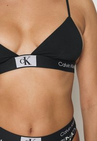 Čierny trojuholníkový bralet s nastaviteľnými ramienkami a širokým elastickým pásom, na ktorom sa nachádzajú biele logá "Calvin Klein" a "CK". Hladká textura.