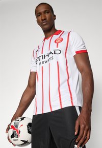 Camisola de futebol branca com riscas verticais vermelhas, gola vermelha e logótipo do patrocinador a preto. Jogador segura uma bola de futebol preta e branca com um logótipo.