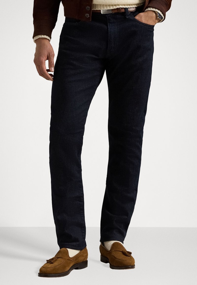Temno modri jeans iz denima s prilega zasnovo, ki vključujejo klasičen stil s petimi žepi in diskretnimi detajli v šivanju. V kombinaciji s prostorskimi rjavimi zamskimi čevlji.