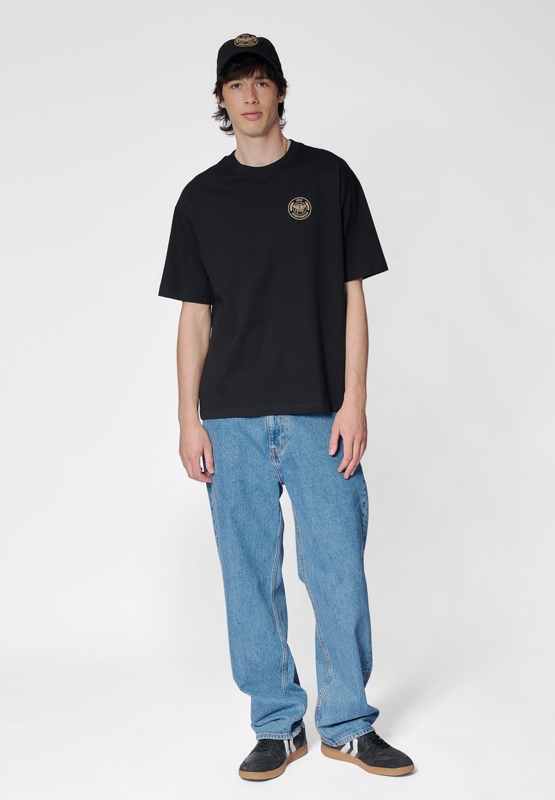 Svart bomullströja med cirkulärt logotyp på bröstet, tillsammans med ljusblå denimjeans och svarta sneakers med mörkgrå detaljer. Kappen matchar tröjan.