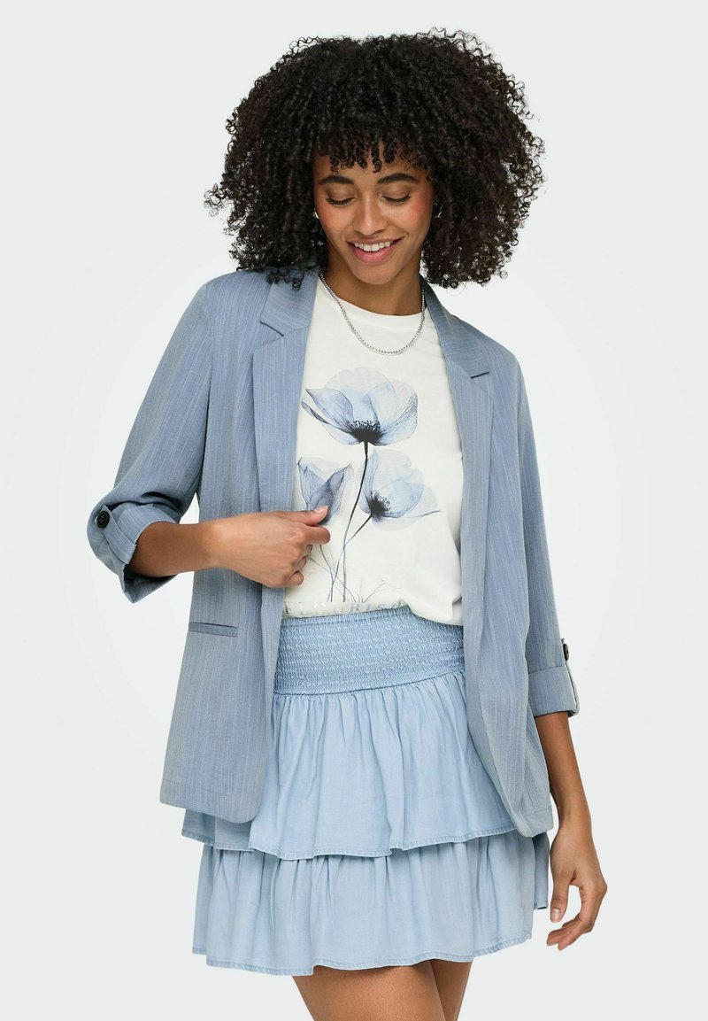 Žena s kudrnatými vlasy, oblečená v světle modrém saku, bílem tričku s květinovým potiskem a vícevrstvou světle modrou sukní, usmívající se a dívající se dolů.