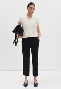 Lichtbeige, geribbelde, korte mouwen gebreide top met een V-hals. Gepaard met zwarte, rechte broek. Model houdt een zwarte handtas vast. Zwarte schoenen.