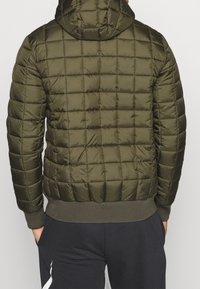 Man in een olijfgroene gewatteerde pufferjack met capuchon, gezien van achteren, gecombineerd met zwarte broeken met een witte streep.