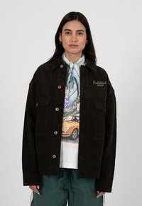 Frau mit glattem, dunklem Haar, die eine dunkelbraune Cordjacke, einen hellblauen gemusterten Schal, ein T-Shirt mit Auto-Grafik und grüne Hosen trägt.