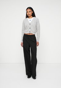 Tânără purtând un cardigan gri scurt, un top alb, pantaloni negri largi și pantofi negri, stând pe un fundal uniform.