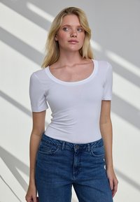 Blondynka w dopasowanym białym topie z krótkim rękawem i niebieskich dżinsach, stojąca na tle białej ściany z ukośnymi cieniami.