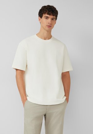 Młody mężczyzna o kręconych ciemnych włosach, ubrany w teksturowaną, jasno-kremową koszulę z krótkim rękawem i jasnobeżowe spodnie, z rękami w kieszeniach, na neutralnym tle.