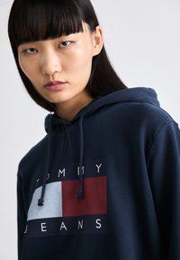 Felpa blu navy in cotone misto con grafica del logo "TOMMY JEANS". Presenta un design rettangolare in rosso e azzurro. Cappuccio con cordino regolabile.