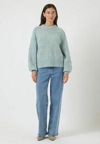 Jasnoniebieski sweter z dzianiny z bufiastymi rękawami i abstrakcyjną teksturą, zestawiony z jeansami o wysokim stanie i beżowymi butami.