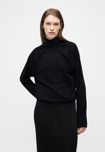 Γυναίκα με ίσια καστανά μαλλιά φοράει μαύρο πουλόβερ με στρώσεις και ψηλό λαιμό και μαύρη φούστα, μπροστά σε απλό λευκό φόντο.